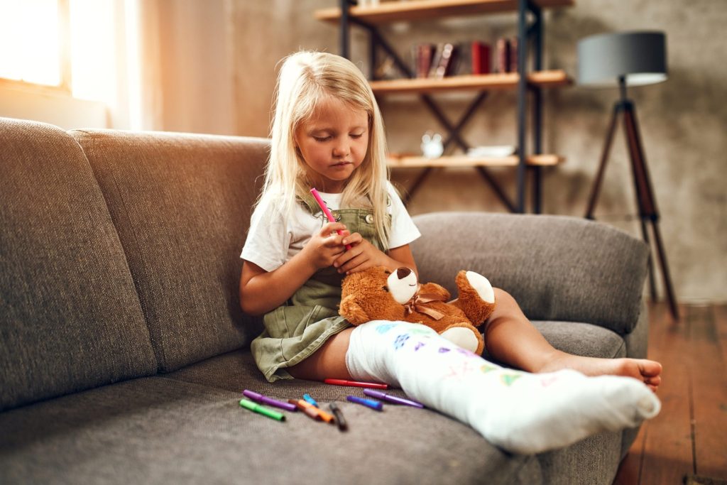 little girl with cast on her leg sitting on the couch with her teddy bear