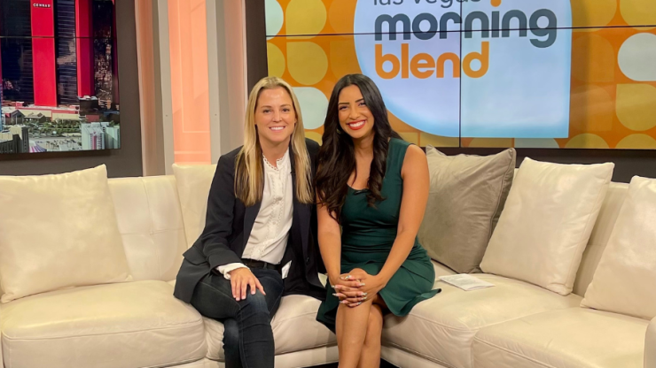 Two confident women share a warm moment on a talk show set, exuding professionalism and care.
