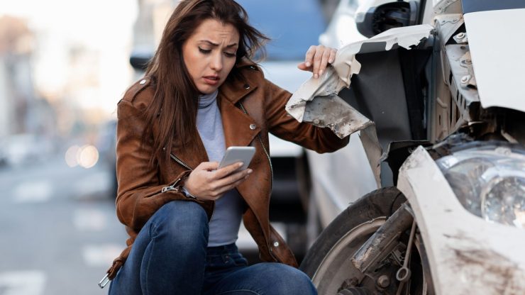 A concerned woman assesses vehicle damage while using her phone, reflecting a moment of urgency.