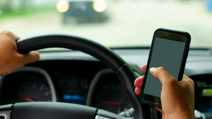 A driver's hand grips the steering wheel while texting, highlighting the dangers of distracted driving.