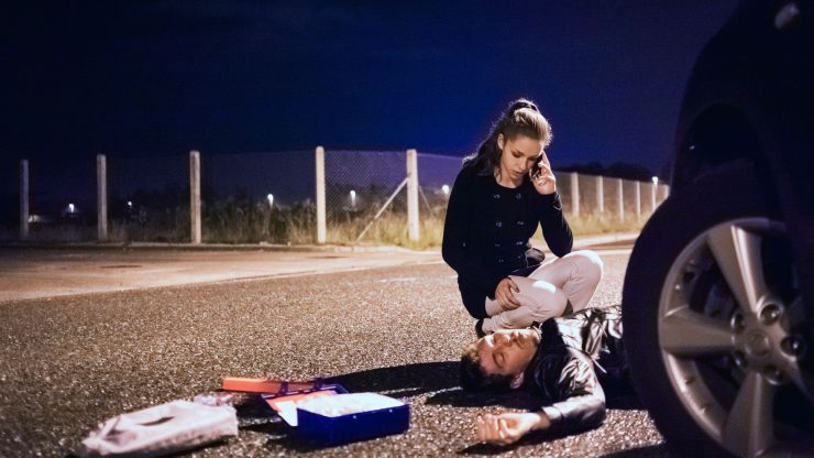 A woman urgently assists an injured individual on a dimly lit road, conveying compassion and urgency.