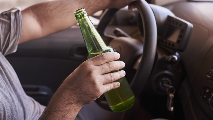 A driver holds a green bottle while steering, highlighting the risks of distracted driving.