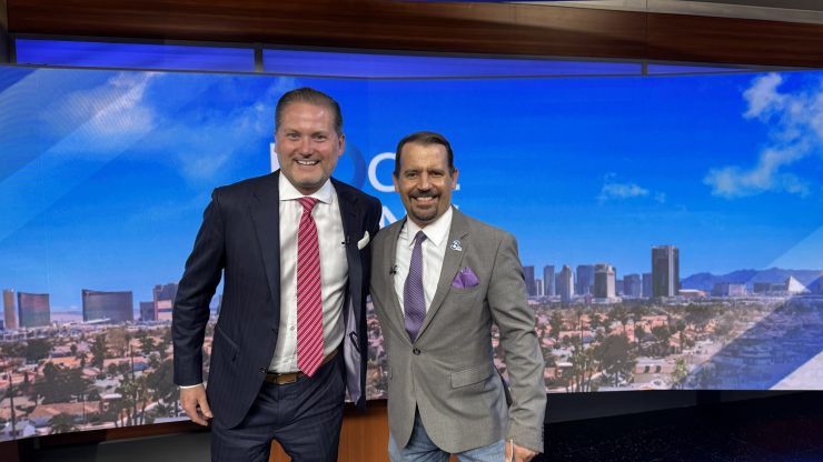 Two confident, professional men in suits stand smiling in a modern studio with a cityscape backdrop.