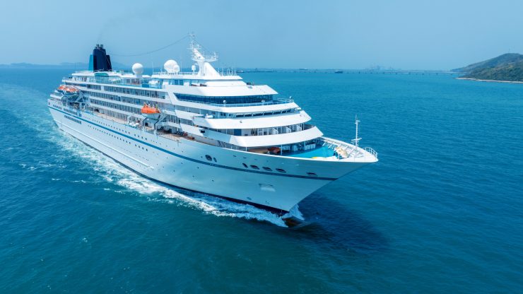 White cruise ship navigating calm blue waters near a coastal shore with clear skies.