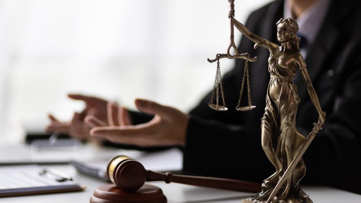 Justice statue and gavel on desk, with lawyer’s hands in blurred background, symbolizing advocacy.