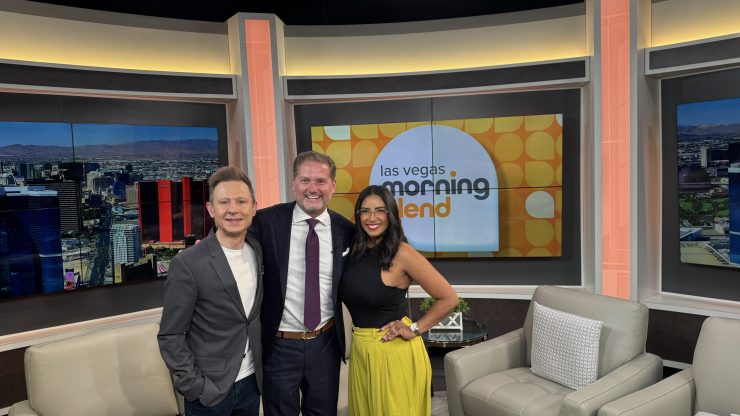 Three professionals confidently pose together in a modern TV studio set with Las Vegas skyline views.