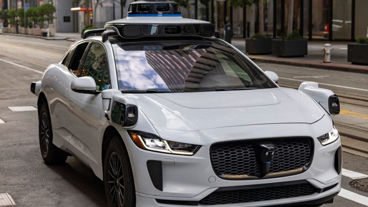 A white autonomous vehicle navigating a city street, reflecting urban architecture in daylight.
